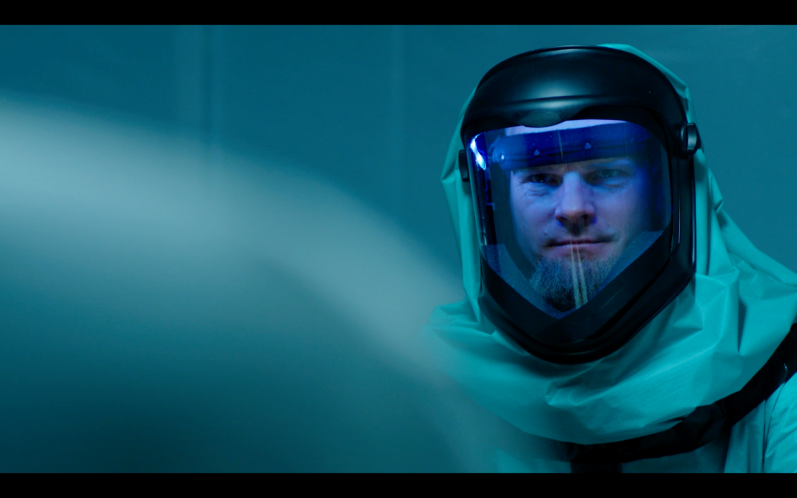 KONTAMINIERT SHORT (2014) Mann in Schutzanzug mit Helm, ernster Blick, blauer Hintergrund.