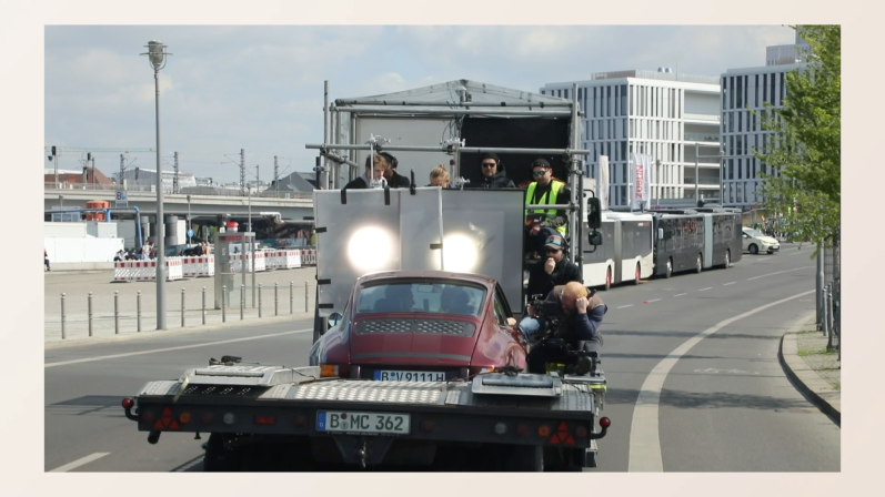 XIAOMI SHORT (2022) Rotes Auto auf einem Anhänger, mehrere Personen auf dem Fahrzeug, Stadtlandschaft im Hintergrund.